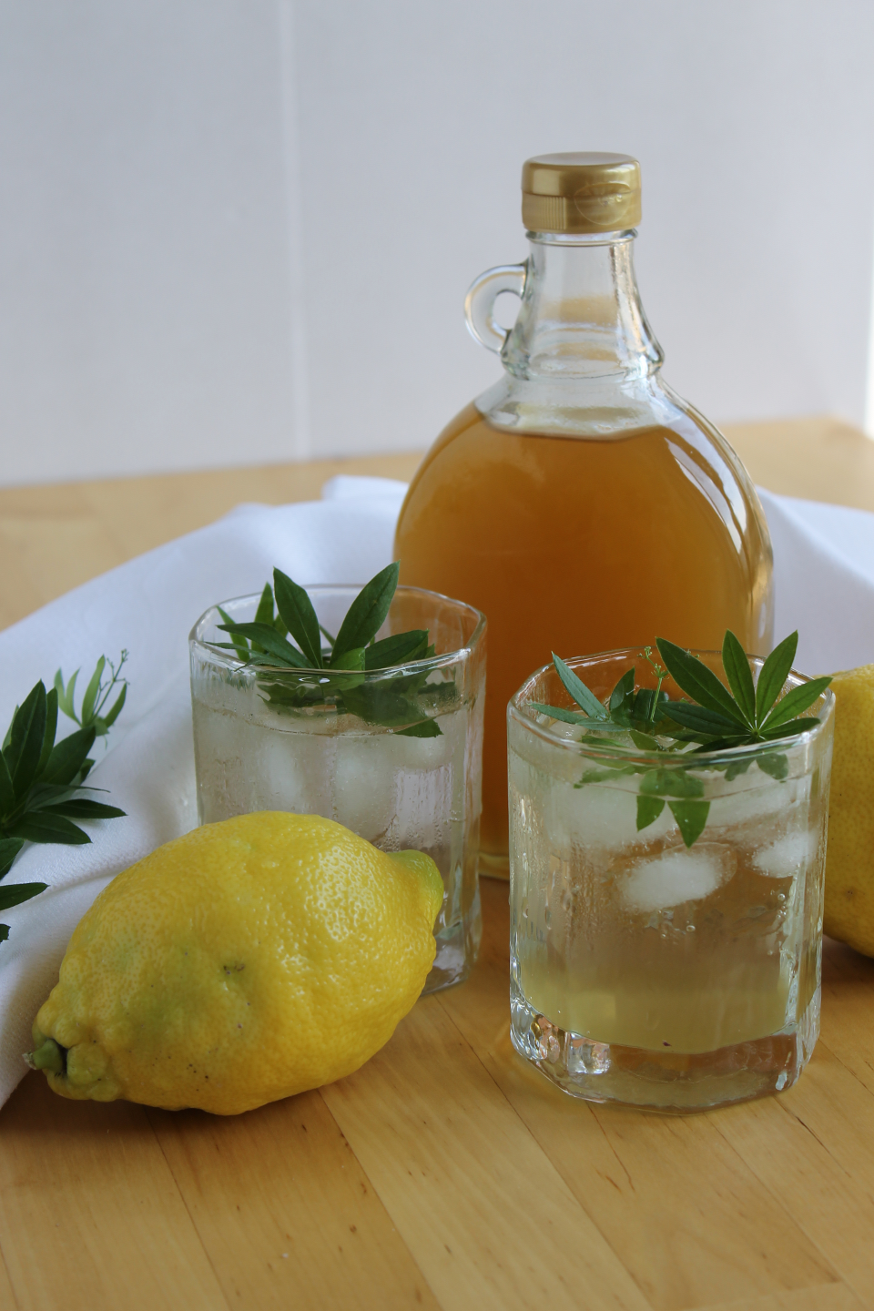 Waldmeistersirup mit Honig in Flasche, davor zwei Gläser mit Waldmeisterlimonade und Waldmeisterblätter, daneben ganze Zitronen und Waldmeisterstängel